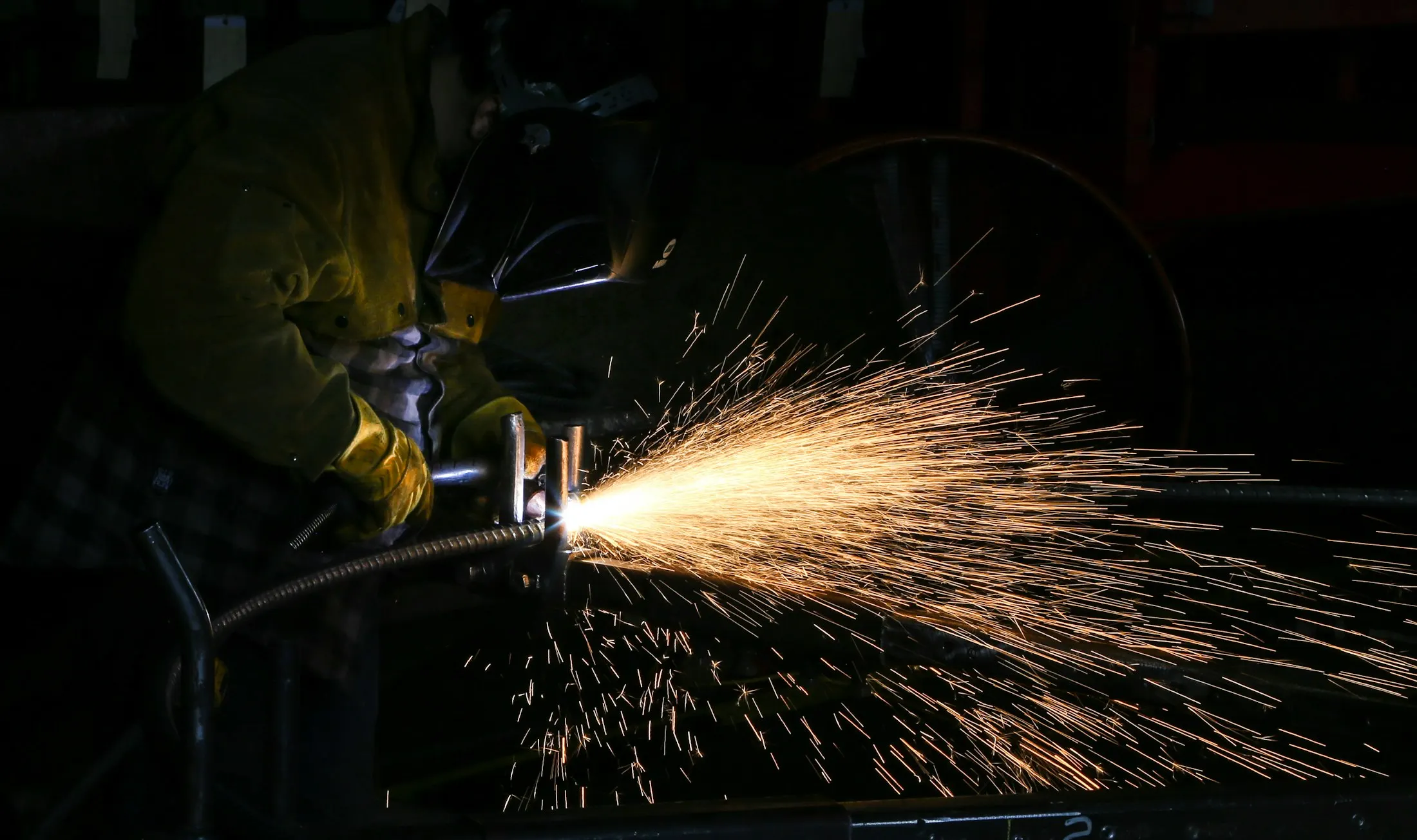 A welder laying a bead on structural steel, molten sparks spraying into the dark of the workshop.